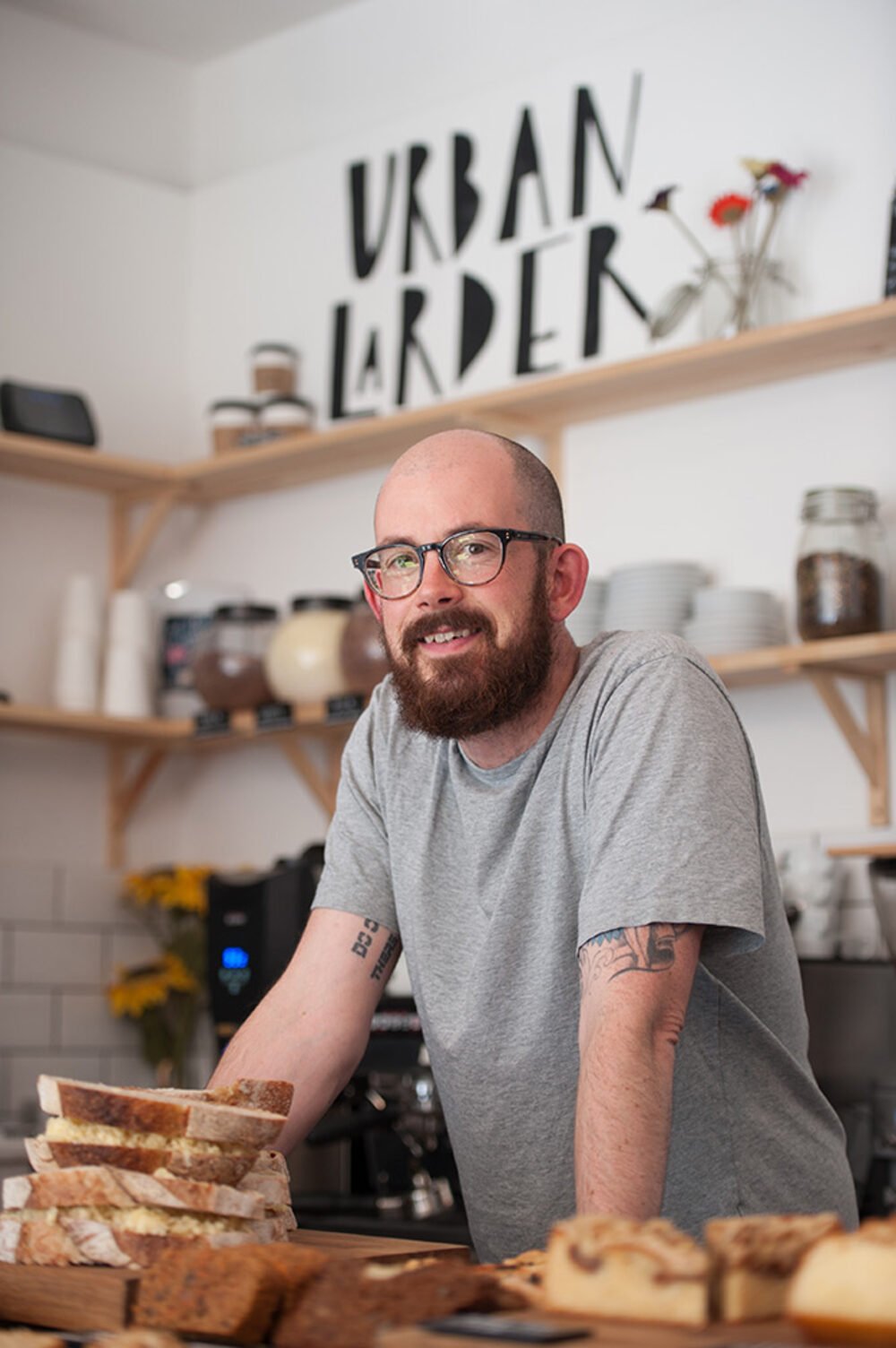The Urban Larder, Cambridge — owner behind the counter with the brand mark on the wall.