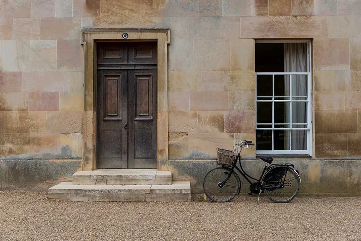 A black bicycle with a wicker basket resting against the pale sandstone wall of Downing College, Cambridge, beside a dark wooden door — stock photography by Jean-Luc Benazet.