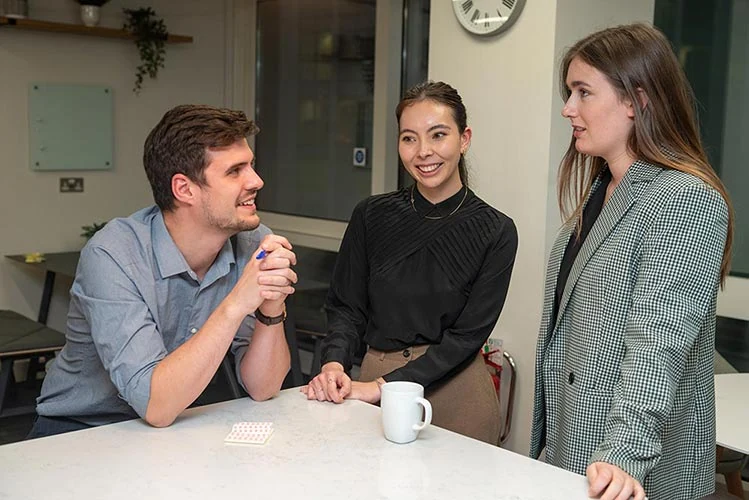 Three people chatting around a counter with mugs in an office