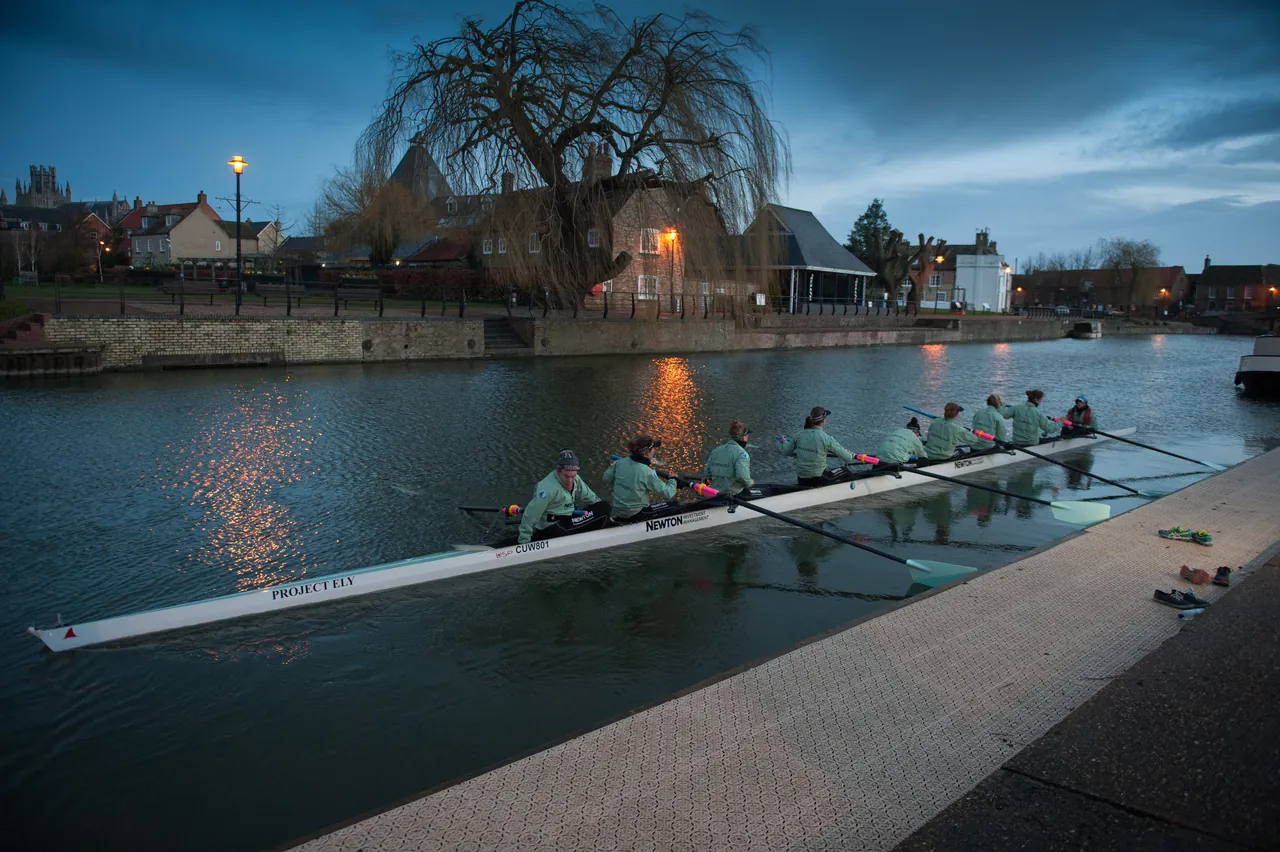 A women’s eight rowing on the River Cam at dusk, with lights reflected in the water