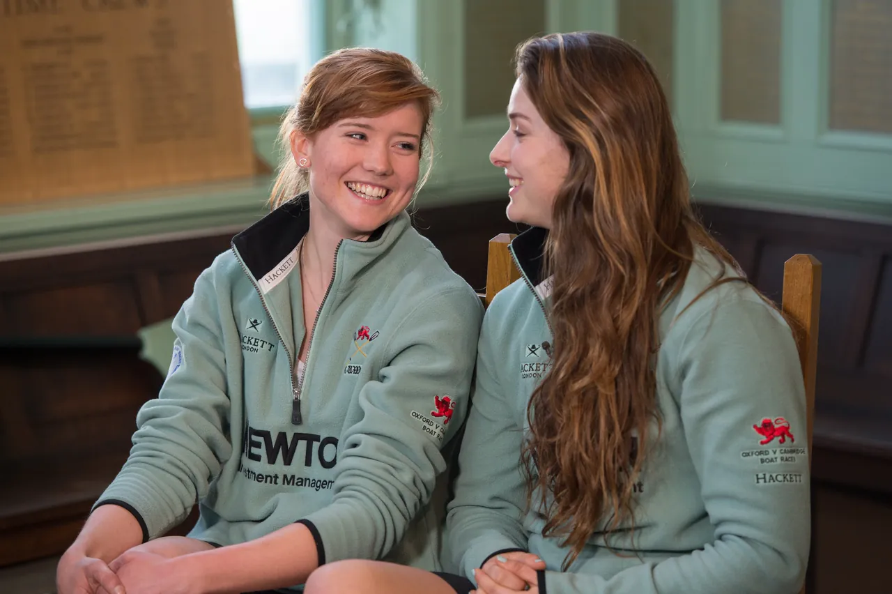 Two rowers in team jackets laughing together in a panelled boathouse room