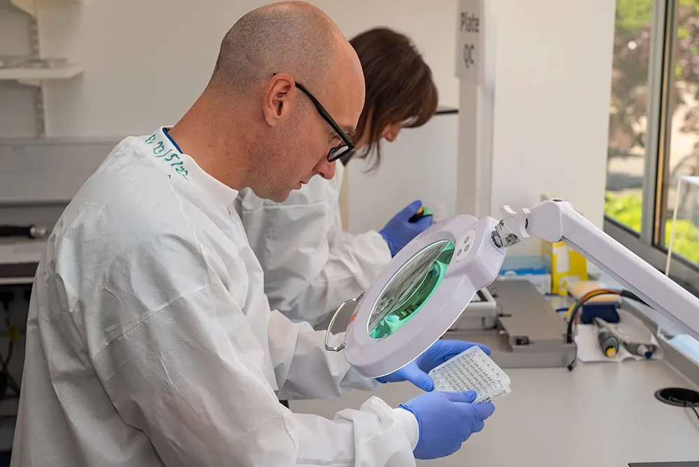 Two scientists in lab coats examining samples under an illuminated magnifier