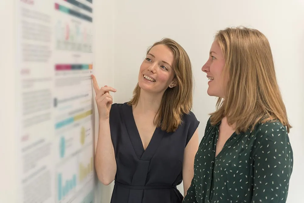 Two women discussing a printed chart on an office wall