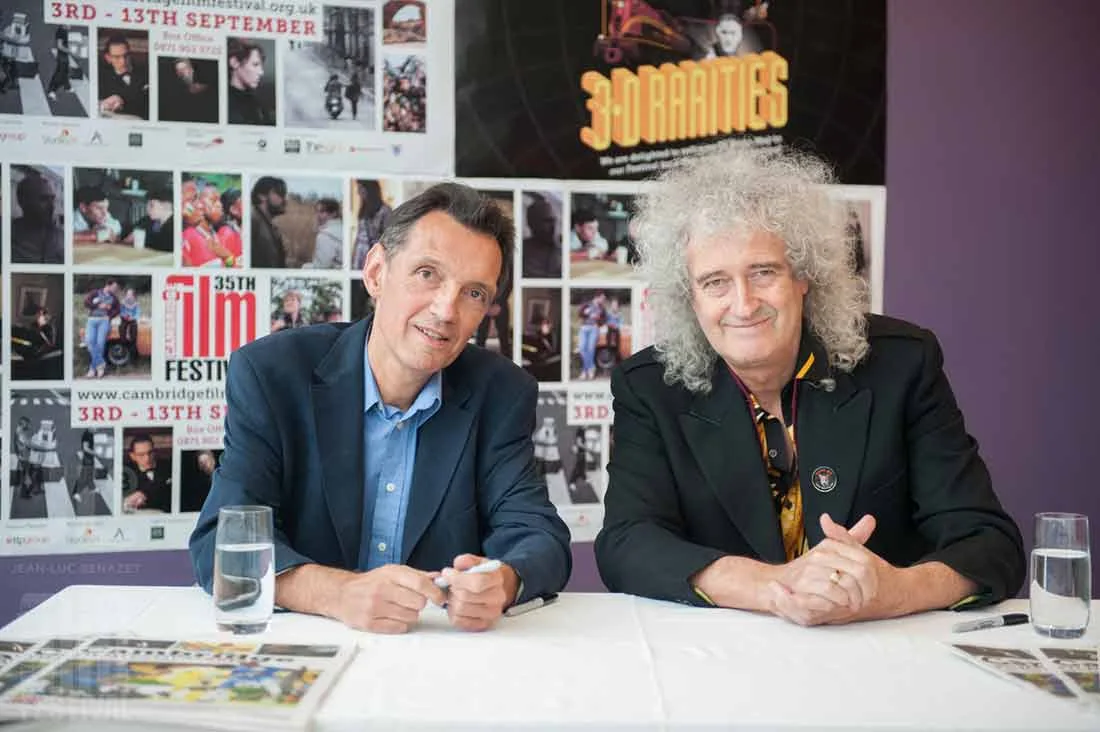 Brian May signing alongside a guest at a table in front of Cambridge Film Festival posters