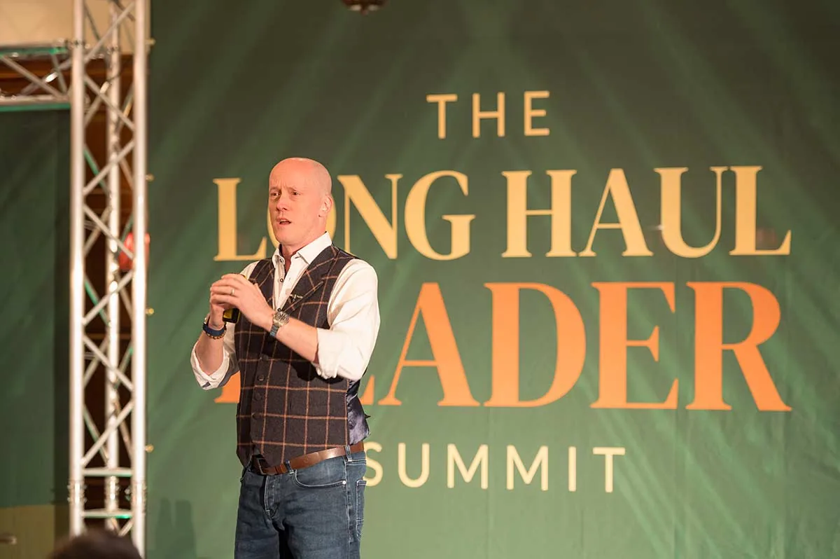 A speaker on stage in front of a large green Long Haul Leader Summit backdrop