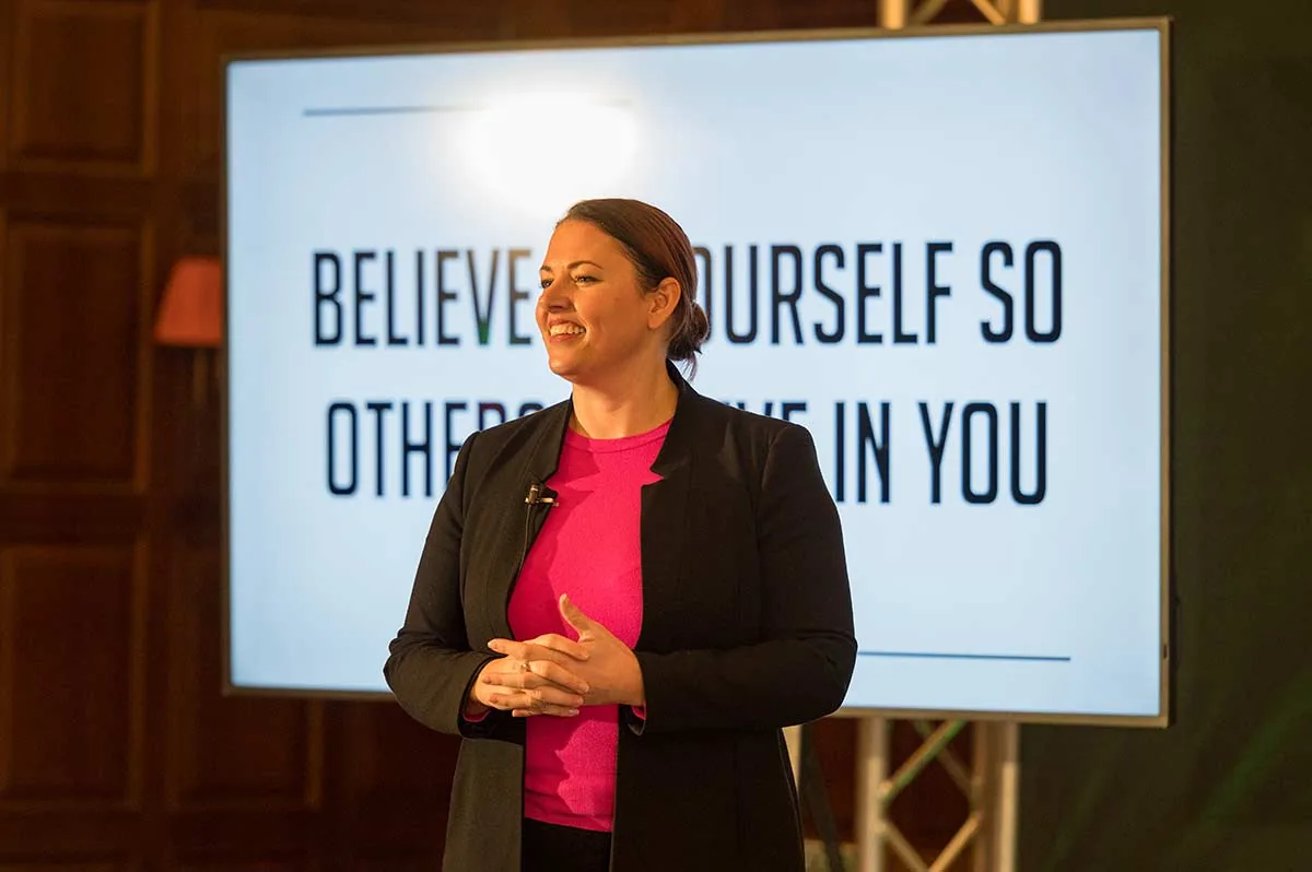 A speaker in a pink top on stage in front of a screen reading Believe in yourself