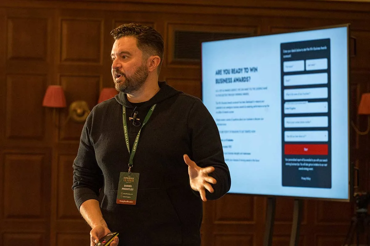 Daniel Priestley speaking beside a screen about business awards in a wood-panelled room