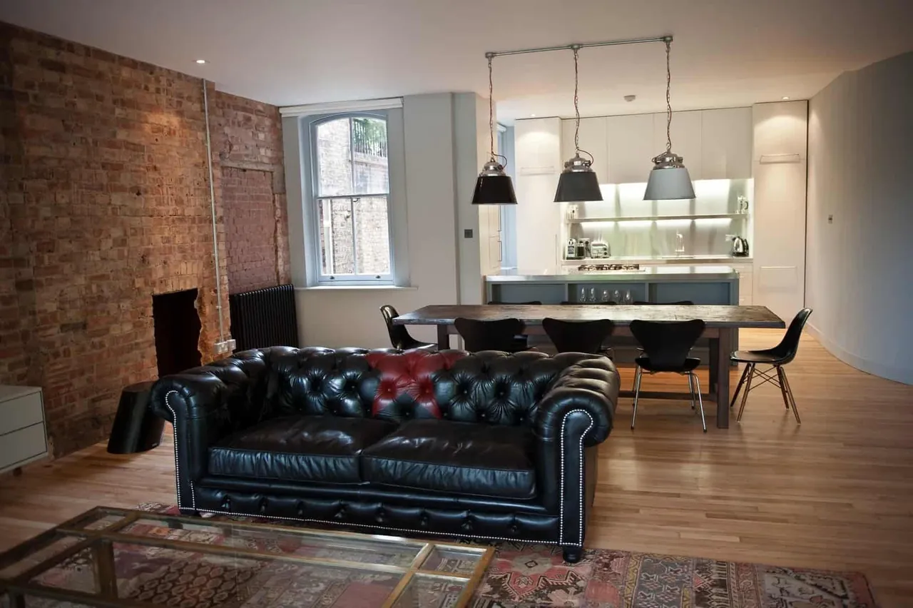 An open-plan loft with a leather sofa, brick walls and a kitchen beyond