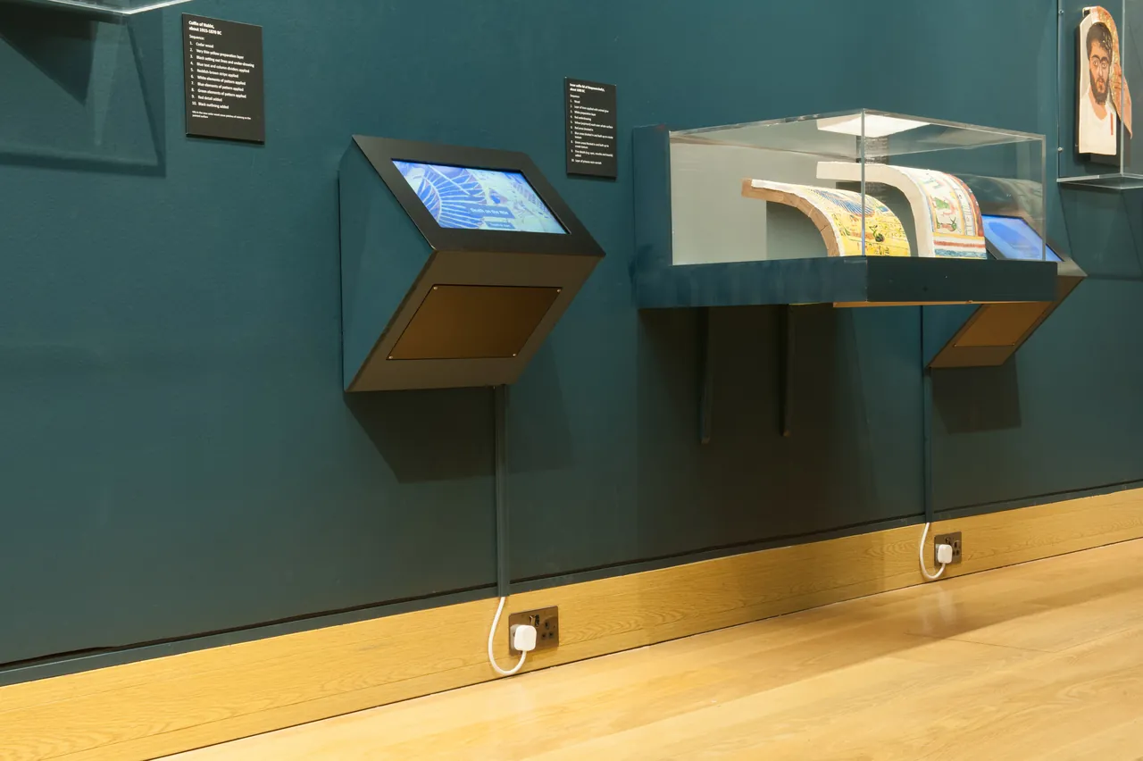 Museum display cases with small screens along a dark teal wall