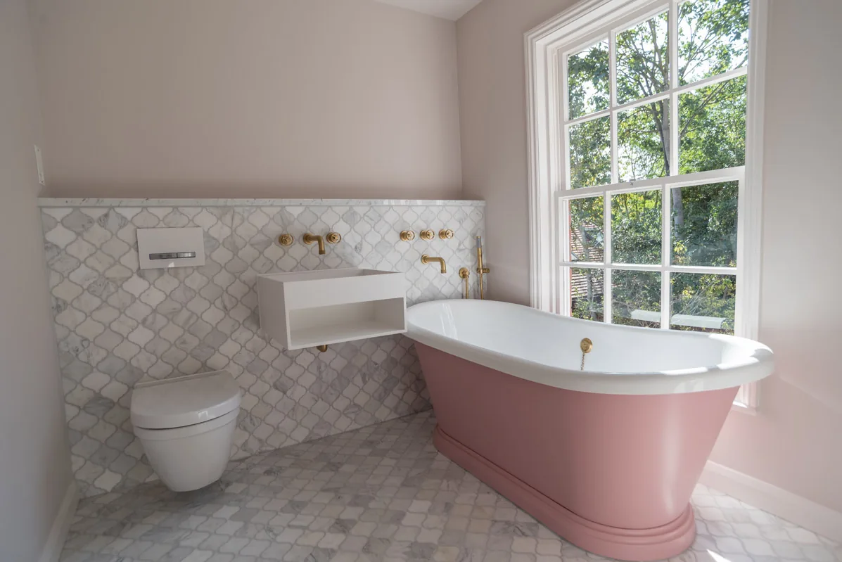 A pink freestanding bath beside a sash window in a bright bathroom