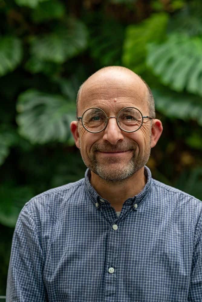 Portrait of a mature man with glasses, smiling, in front of lush green foliage.