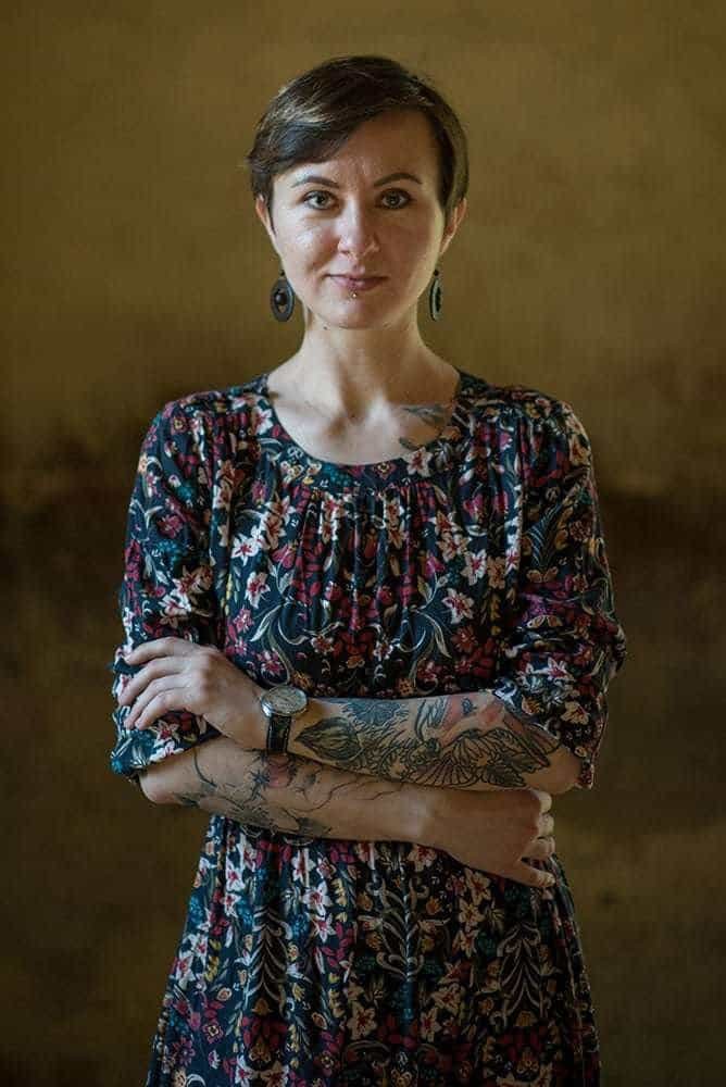 Portrait of a confident woman with tattoos, wearing a floral dress, against a warm background.