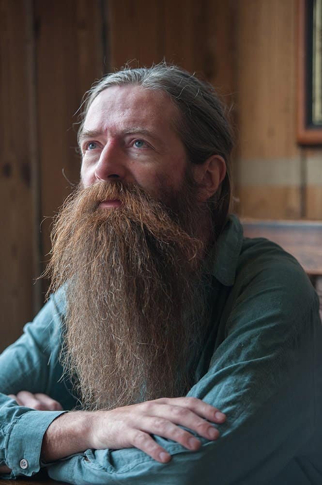 Portrait of a bearded man with long hair, sitting indoors with natural light.